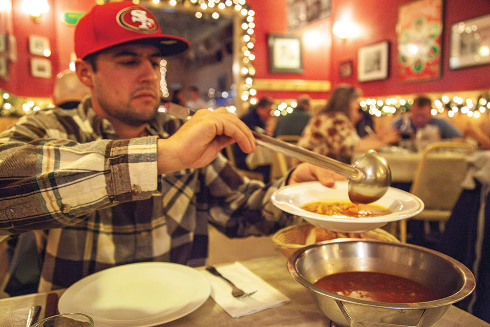 A man in a hat ladles soup into his bowl from a larger bowl sitting on the table. The restaurant is decorated for Christmas.