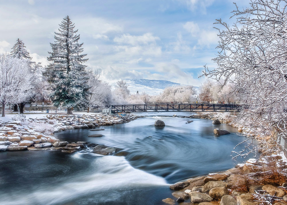 Icy blue Truckee River with a bridge going across and snowy rocks and trees on the shore. Snowy mountains and clouds in the distance.