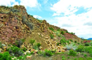 Stone wall crumbles under blue sky, surrounded by green vegetation and scattered rocks in a rugged landscape.