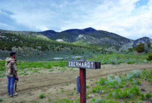 Signpost reading "EBERHARDT" stands in green landscape with mountains; person stands nearby, holding an item, looking at scenery.