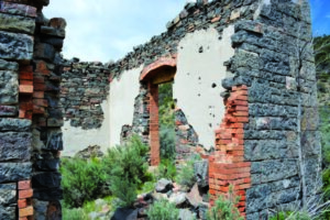 Ruined brick and stone walls stand exposed, surrounded by overgrown bushes and clear blue sky overhead.