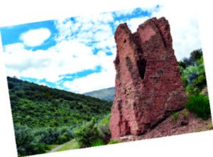 Red rock formation stands upright amidst lush greenery under a bright blue sky with scattered clouds.