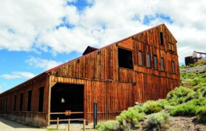 Wooden barn stands abandoned, nestled on a hillside under a partly cloudy blue sky, surrounded by greenery.