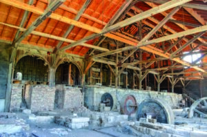 Old machinery remains stationary amidst wooden beams and brick walls inside a large, partially roofed industrial structure.