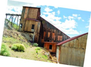 A weathered wooden building stands on a hillside, surrounded by sparse greenery, under a bright, cloudy sky.
