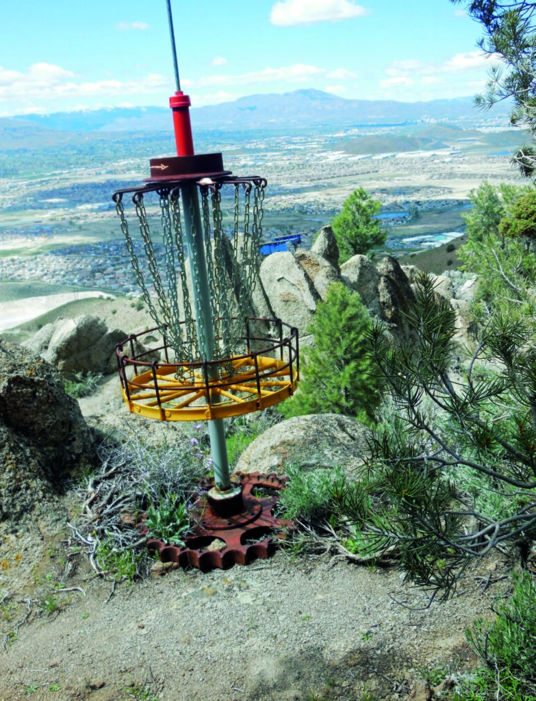 Disc golf basket stands idle on rocky hilltop, surrounded by trees, with sprawling valley and distant mountains in background.