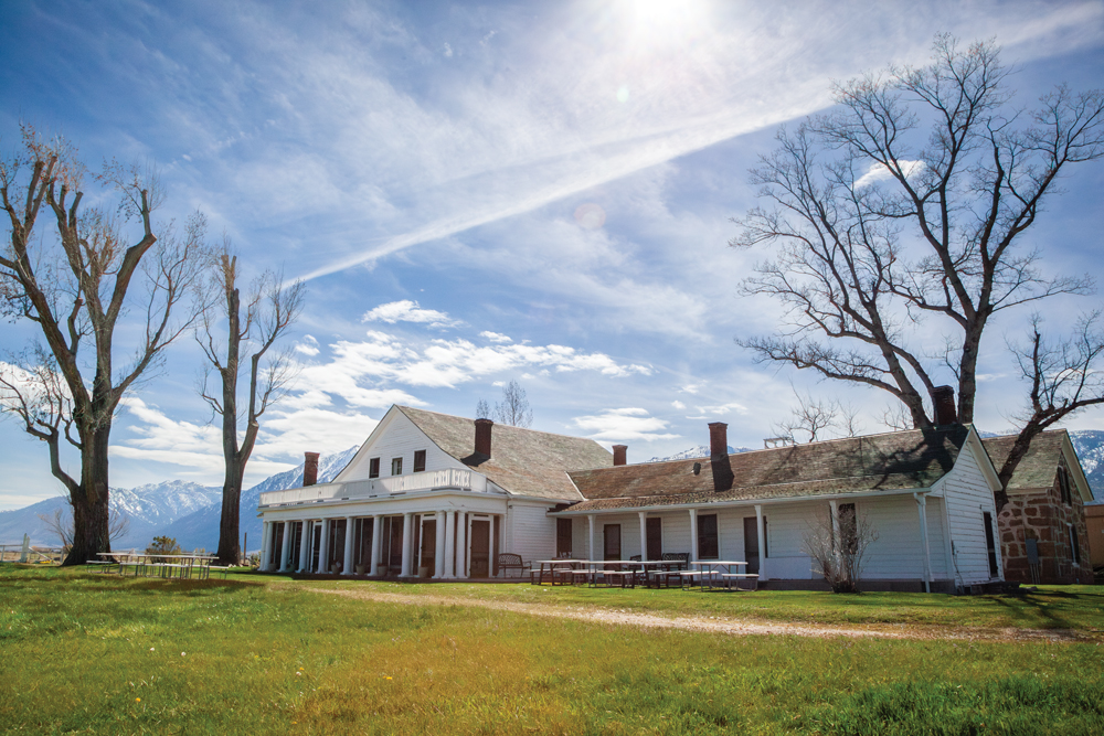 An exterior shot of Dangberg Home Ranch. A white medium sized building sits in front of the Sierra Nevada mountains, which have a light dusting of snow, on a mostly sunny day with clouds lightly filling the sky. Beside the house sits three large, barren trees. Greenish grass stretches out in front of the house with a path dividing the greenery, and tables with bench seats sits on the left of the image and right in front of the building. A second brick building peeps out behind the first building on the right of the photo.