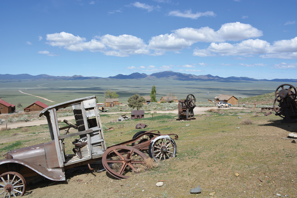 Rusty vintage car sits abandoned in a grassy field, surrounded by old machinery, with mountains on the horizon.