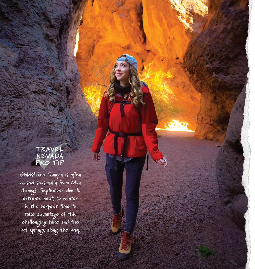 A young blonde woman walks through a red-stone canyon. Behind her it is lit brightly by the sun. She is dressed for cooler weather, with hiking boots and a hat, evoking a sense of wonder and adventure. Copy on the image says: Goldstrike Canyon is often closed seasonally from May through September due to extreme heat, so winter is the perfect time to take advantage of this challenging hike and the hot springs along the way.