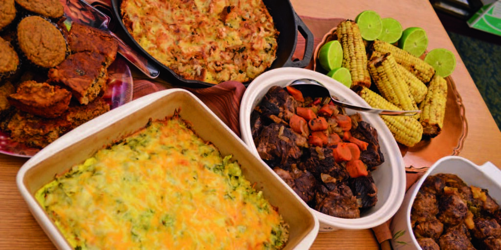 Assorted dishes on a table include roasted corn, meat stew with vegetables, casseroles, and muffins, creating a hearty spread.