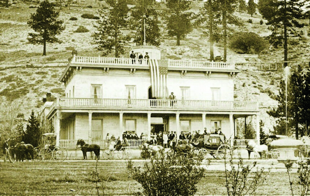 Building with American flag, people standing and horse-drawn carriages in front, set against a wooded hillside background.