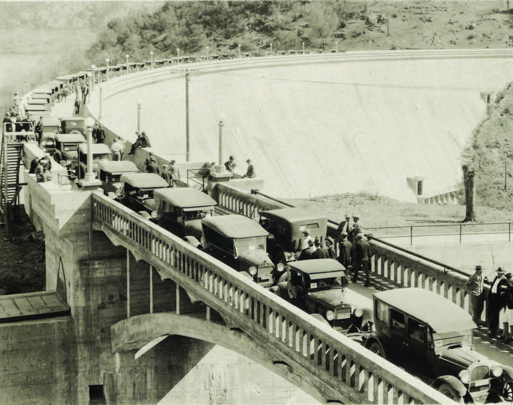Cars are parked on a bridge; people are walking, socializing near a large curved dam structure in the background.