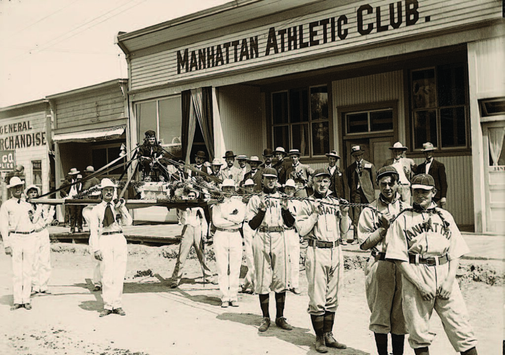 Baseball players carry a costumed person on a litter outside Manhattan Athletic Club; spectators watch nearby. 