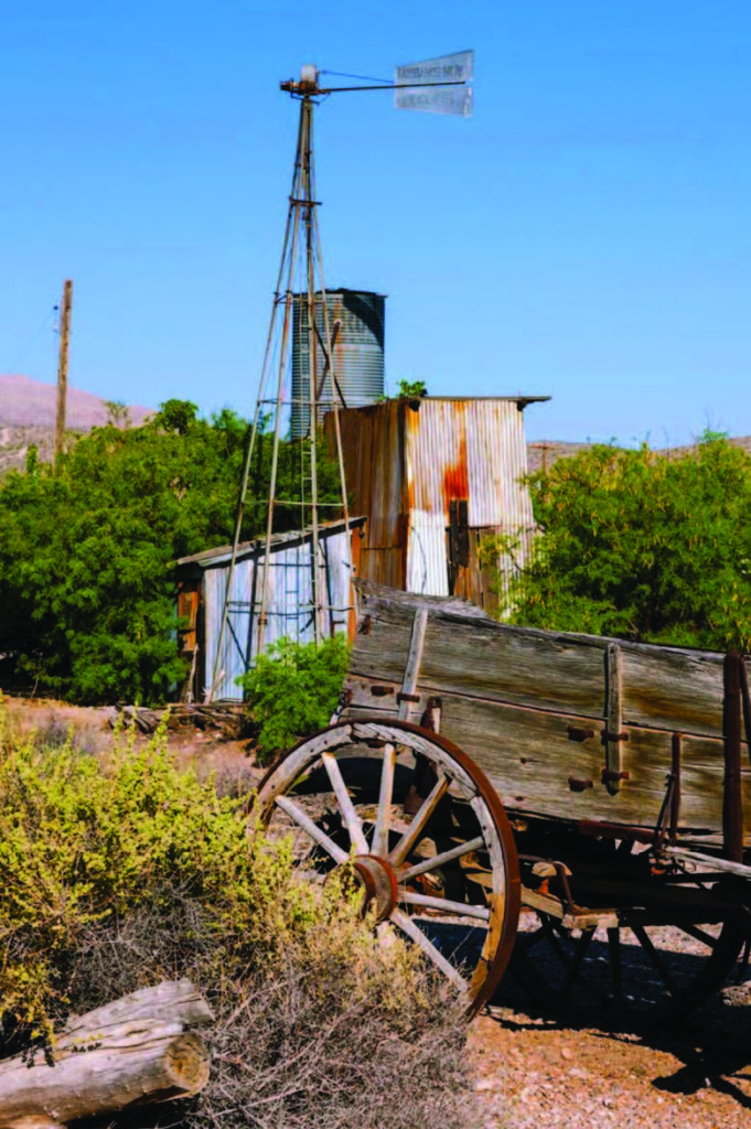 Old wagon rests beside a dilapidated, rusty corrugated metal shed, with a tall windmill nearby and desert vegetation surrounding.