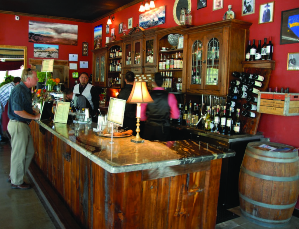 Bartender serves drinks at a rustic wooden bar, surrounded by wine bottles and framed artwork on red walls.