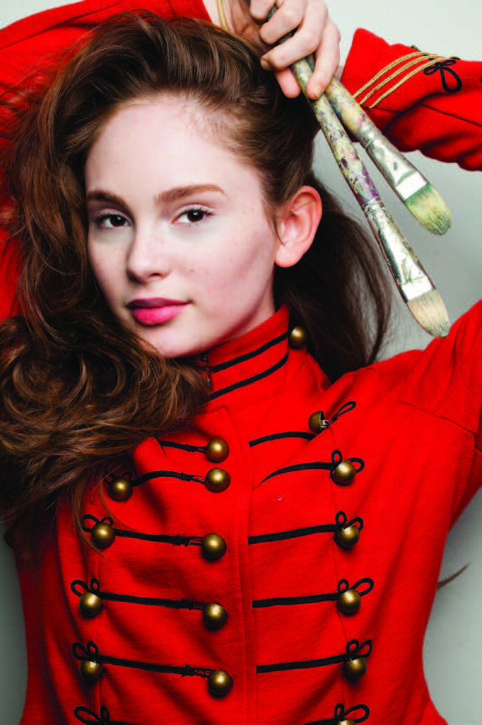 Person holds paintbrushes, wearing a red jacket with gold buttons, against a neutral background.