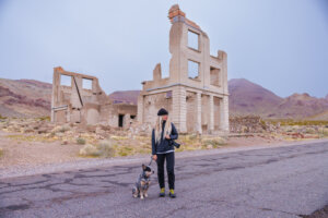 Woman holding a camera, standing with her dog in front of a crumbling building at Rhyolite.
