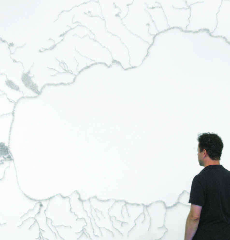 Man observes large, abstract map artwork with intricate river-like lines on a plain wall.