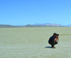 Person squats, photographing in a vast desert landscape under clear blue sky, distant mountains visible at horizon.