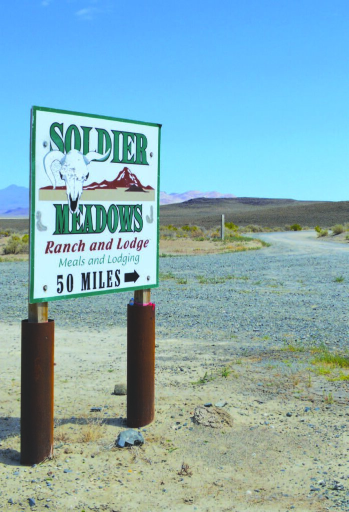 Signpost reads "Soldier Meadows Ranch and Lodge Meals and Lodging 50 Miles" beside a dirt road and open field.