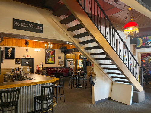Interior photo of Sassafras Eclectic Food Joint shows a wooden bar with black chairs, tables and a wine-colored booth in the dining area, swag lamp with stairway, lots of psychedelic-styled artwork, and a sign that reads "Be nice or leave."