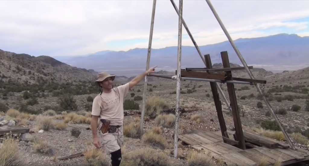 A person points beside a wooden mining frame in a desert landscape with distant mountains and sparse vegetation.