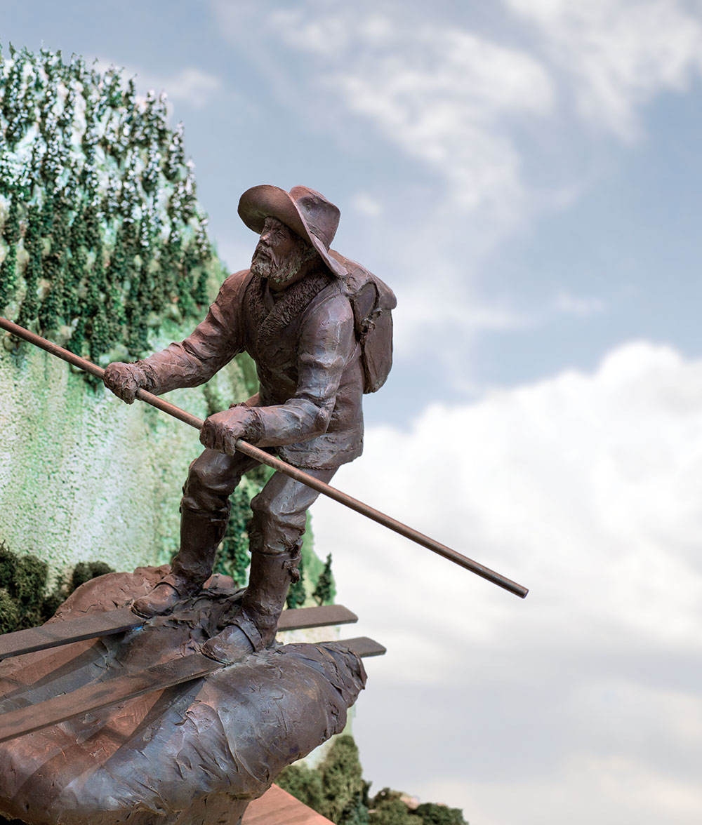 A bronze statue of Snowshoe Thompson on skis, with a modeling of a mountain and trees behind him. Background is blue skies with puffy white clouds.