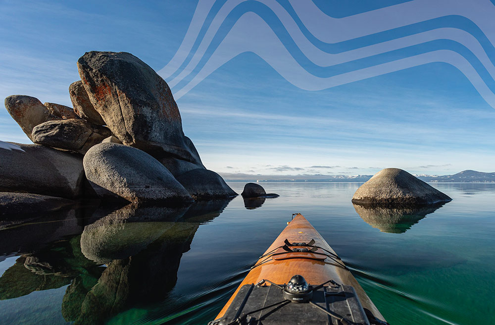 The bow of a wooden kayak drifts through emerald waters, toward an opening in large boulders that are reflected in the lake. There are snow-capped mountains in the distance.