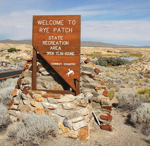 Welcome to Rye Patch State Recreation Area sign. Reads 'open year round, Cowboy Country.' The sign is set in stone, and there is a reservoir, road, desert scrub, and mountains behind. Photo ©Nevada State Parks