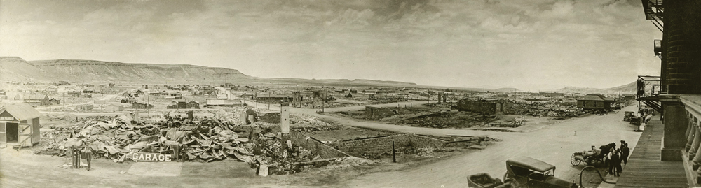 Panorama of Goldfield in 1923 after a large fire wiped out the town. Rubble where buildings once stood sprawl across the town. Roads and telephone poles can be seen amongst the rubble. On the bottom right hand corner of the image, cars and people can be seen standing next to a building.