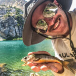 A man wearing a hat and sunglasses, smiling at the camera while holding a fish. A lake is behind him.