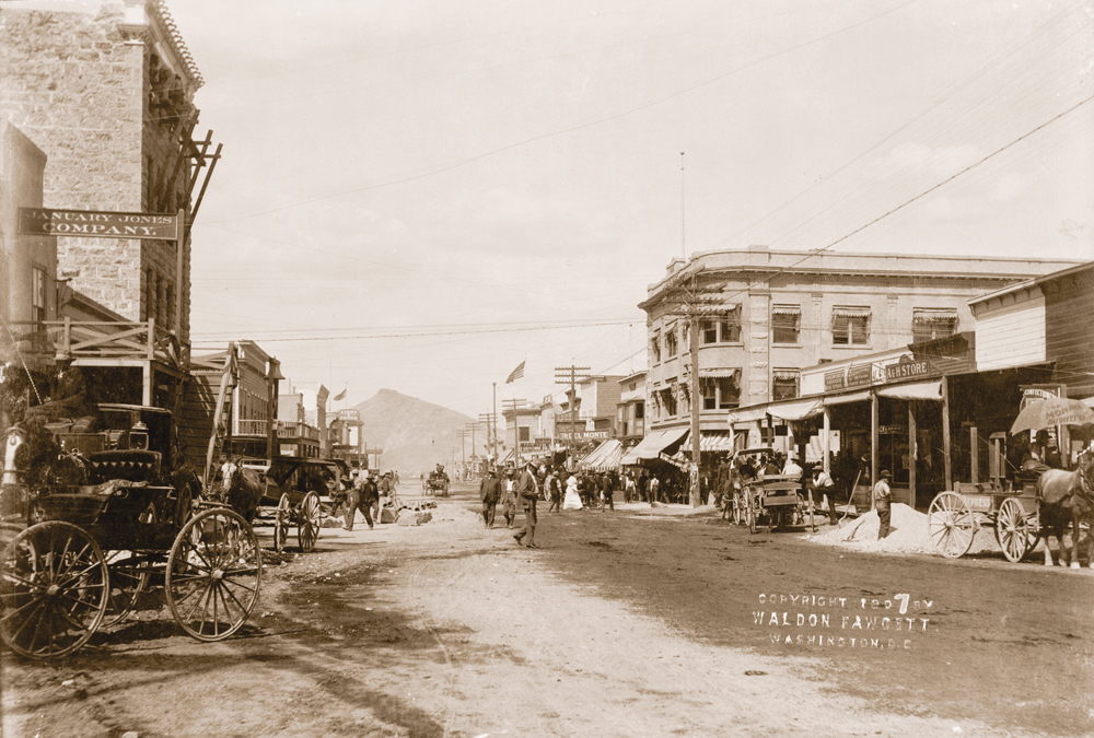 Old historic picture of Goldfield circa 1907. Buildings line a dirt road street as people are walking down and across the street. There are horse drawn carriages parked along the side of the street. Telephone poles and lines tower over the street. In the distance sits a small mountain.
