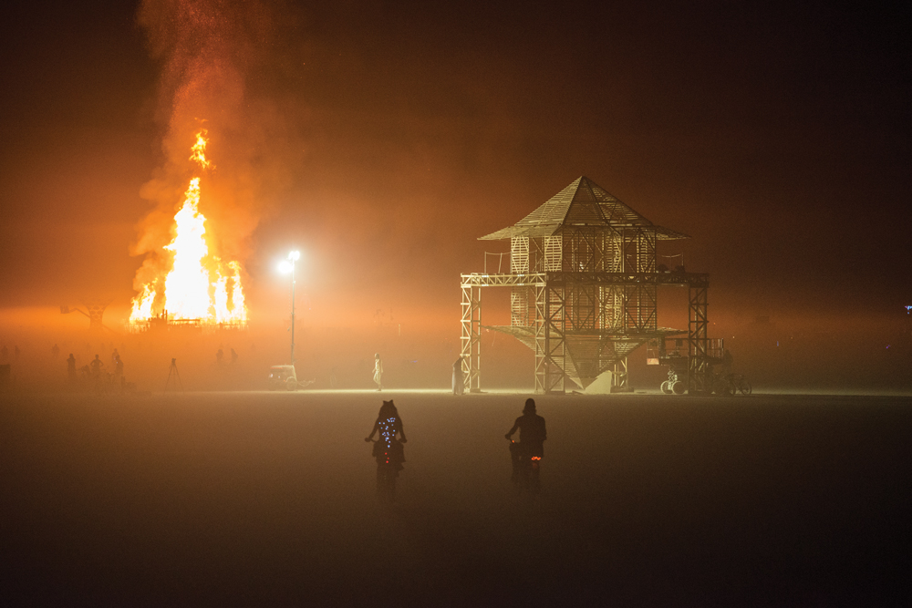 Two people riding bicycles on the playa during Burning Man at night. A structure is on fire in the background.