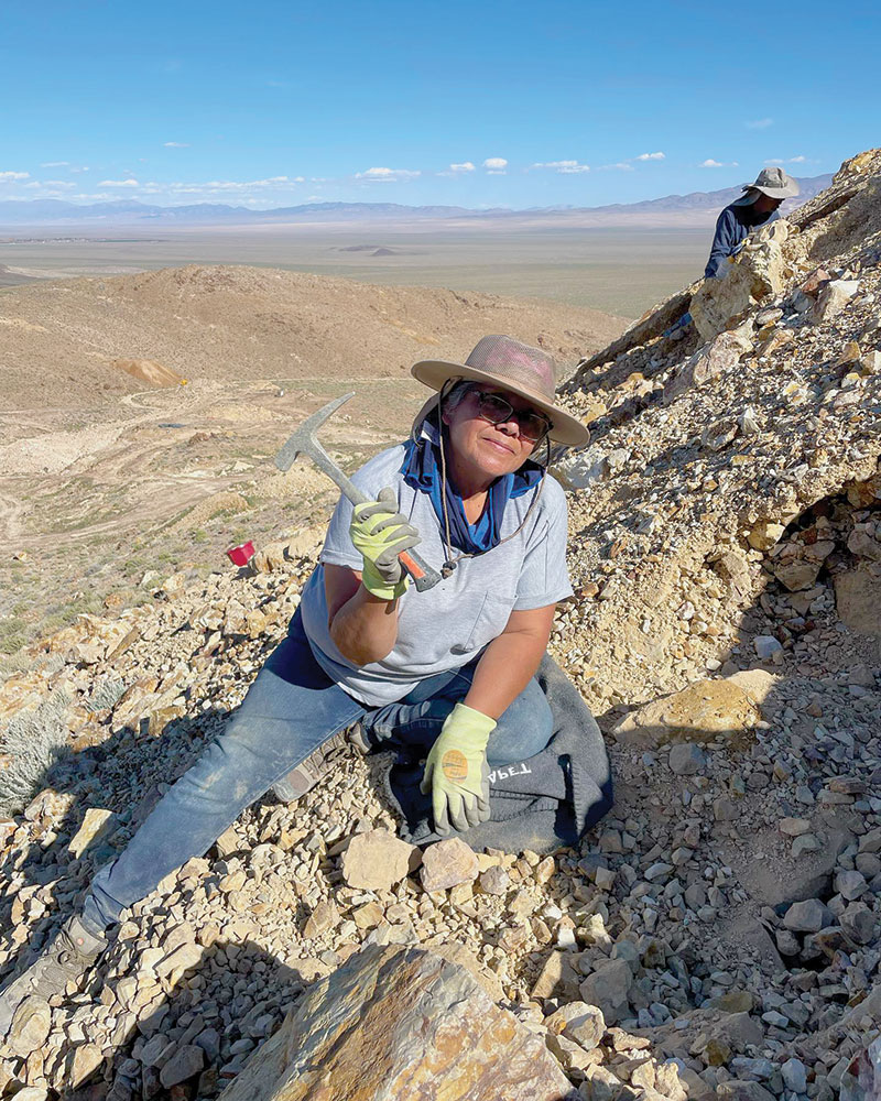 A woman stands on the side of the hill, mining for turquoise with a rock hammer