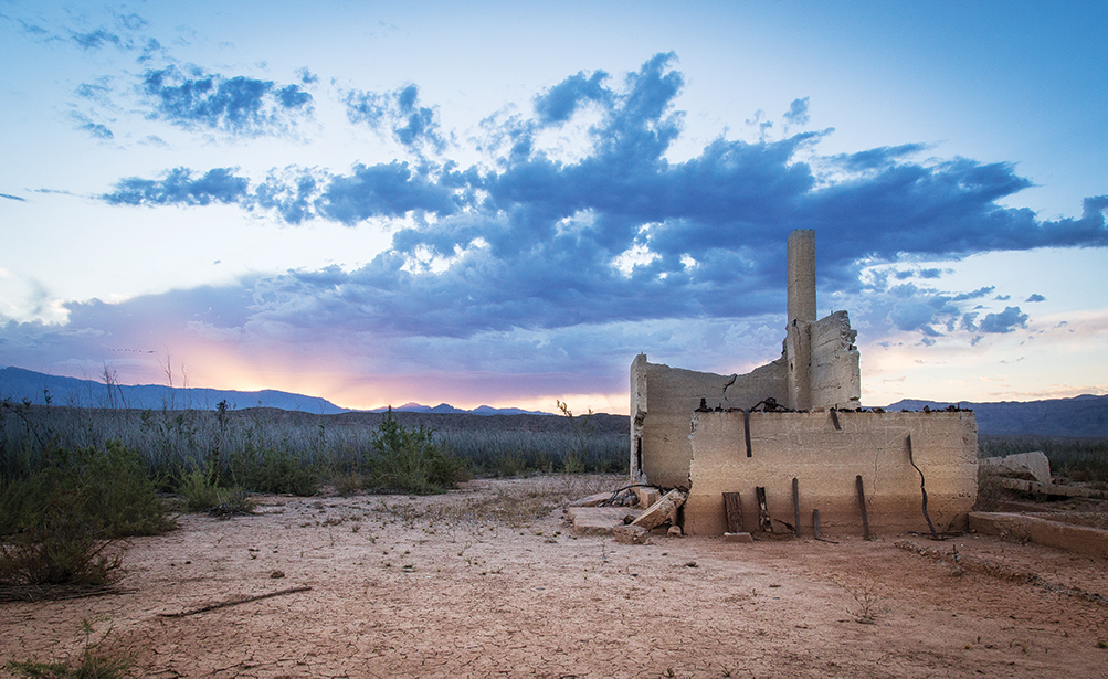 Ruined structure stands abandoned in a barren desert landscape beneath a colorful sunset sky with scattered clouds.