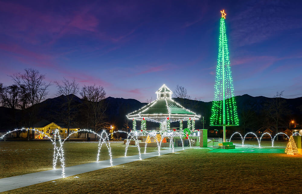 A park is lit up for winter holidays. It is dusk, there are mountains behind the scene, and pink clouds in a dark blue sky.