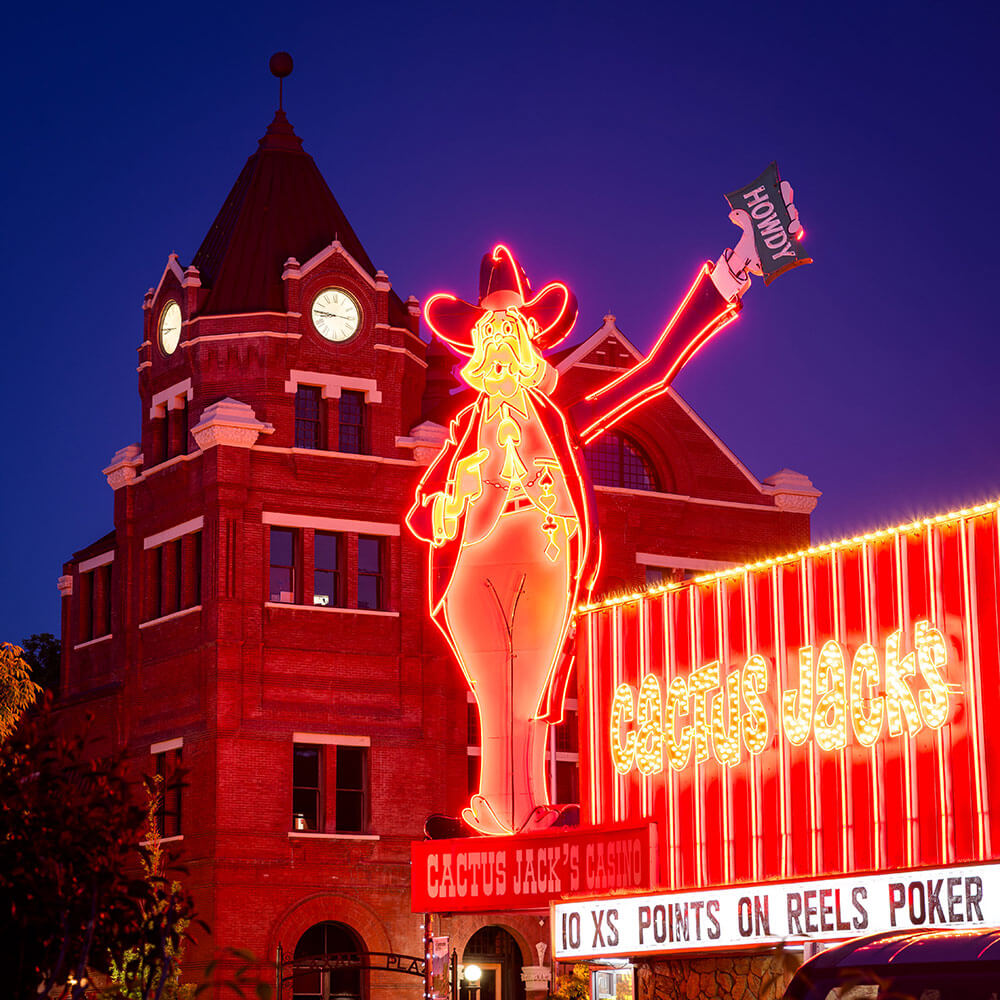 A neon figure waving a sign that reads "howdy." Cactus Jacks Casino, with a historic brick building and clock tower in the background.