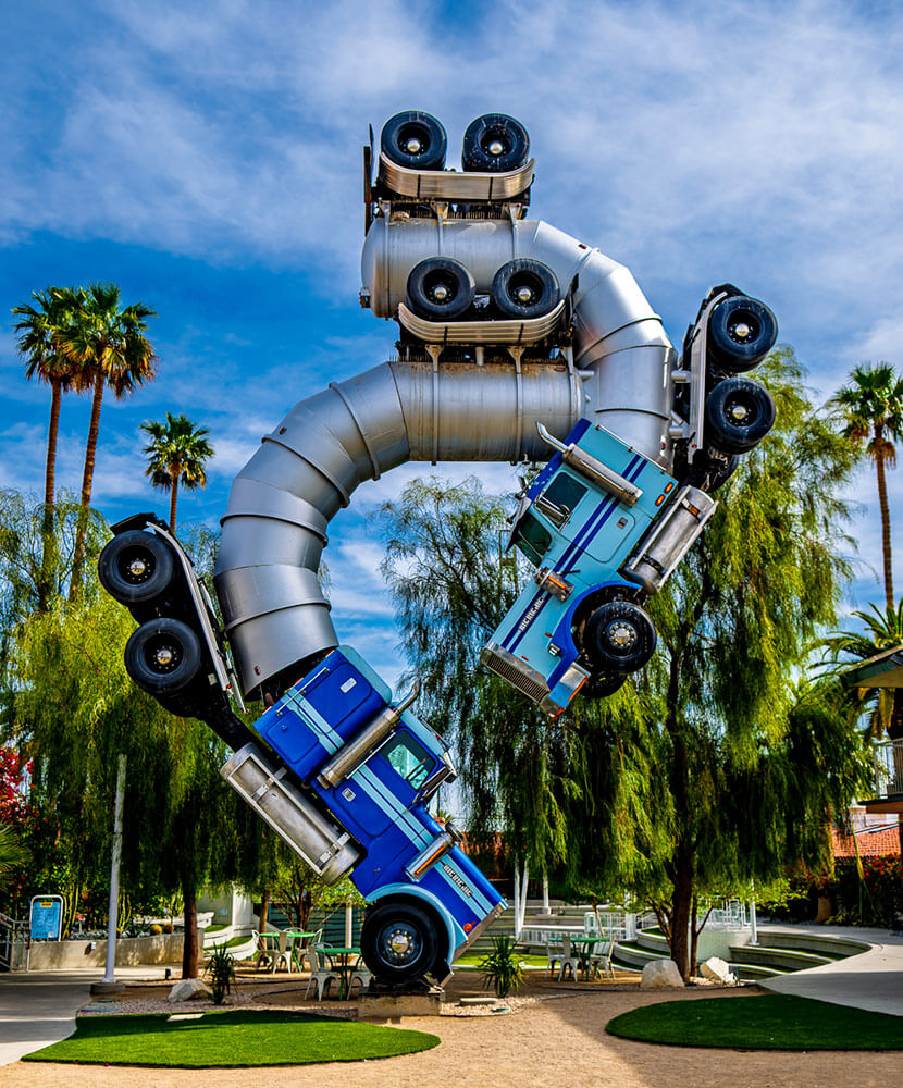 Burning Man art. Two semi trucks with stylized containers on the background, connected and rising into the air, surrounded by palm trees and a park-like setting.