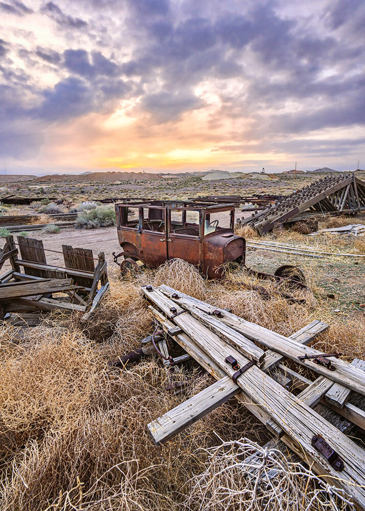 A ghost town setting with old wood and metal on the ground. An old rusted car sits in the middle of all of it, with the sun setting in a cloudy sky.