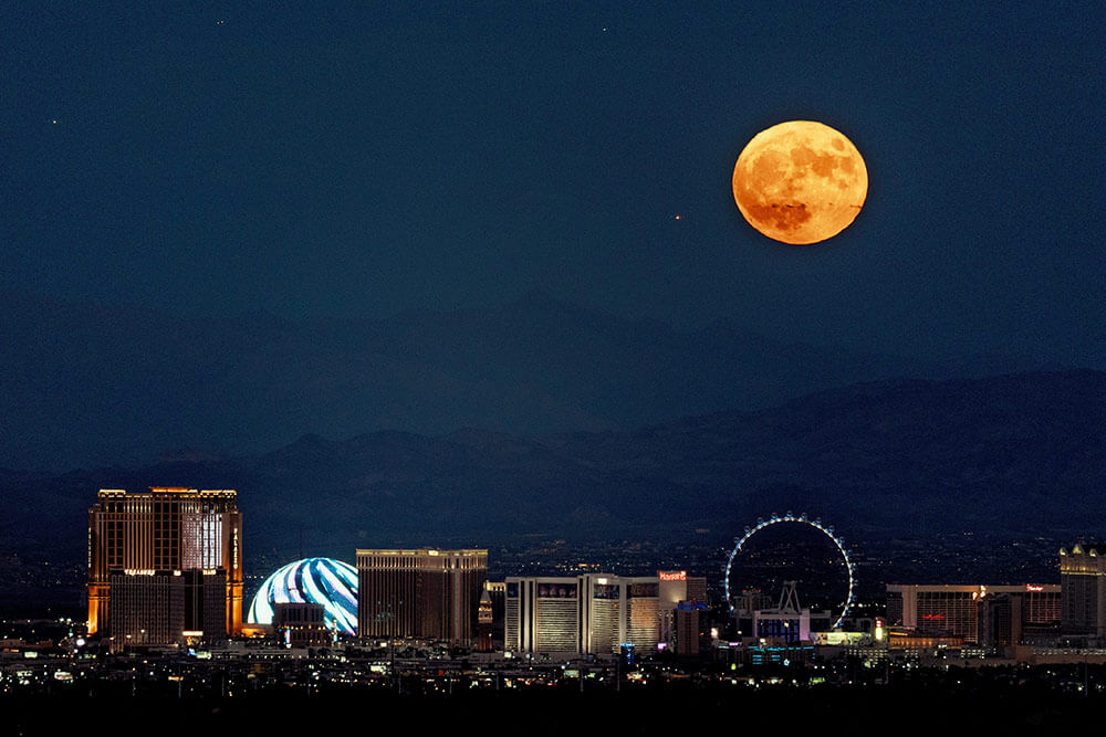 Las Vegas at night, lit up, with mountains in the background and a large orange moon with a few stars in the sky.