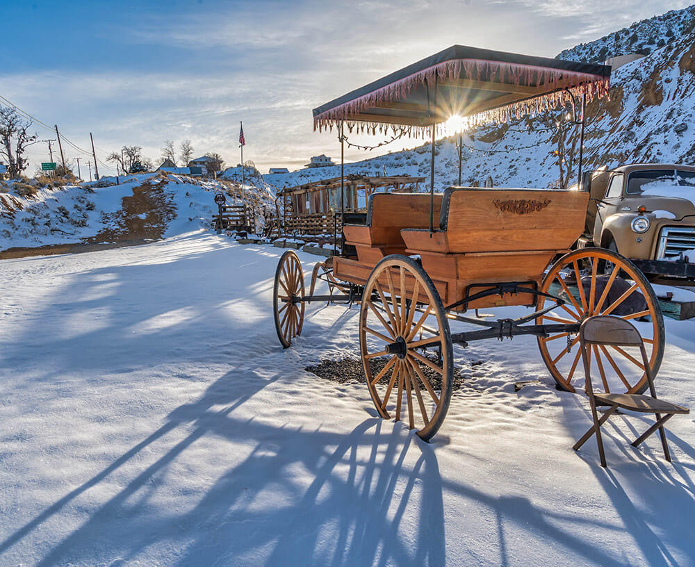 An antique wooden wagon sits in the snow, with other antiques surrounding. There is a sunburst showing, with a cloudy blue sky.