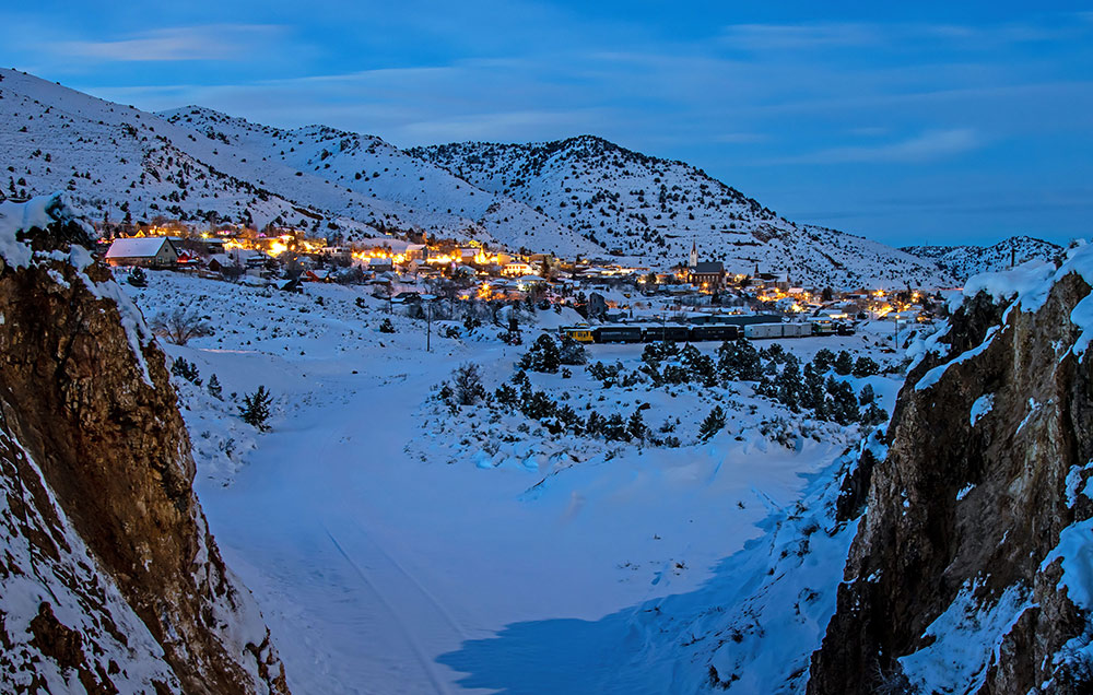 A mountain town lit up at dusk, with snowy mountains surrounding.