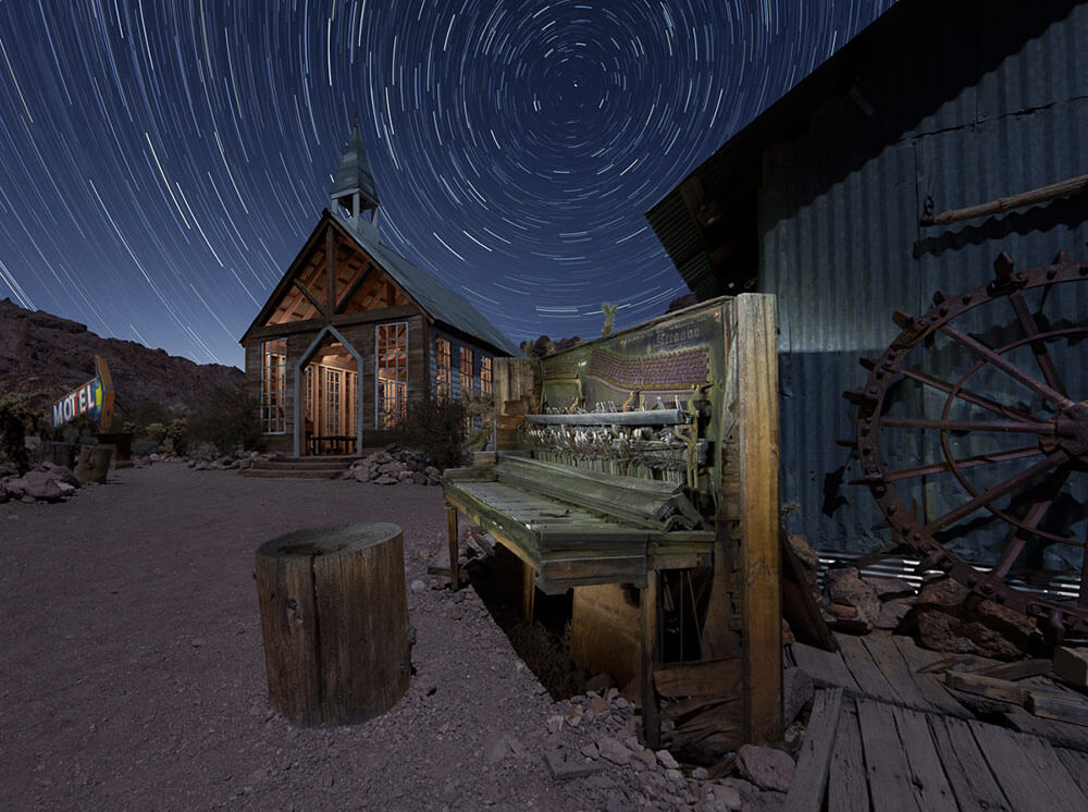 Long exposure shot of a ghost town building with an antique piano in front of it. Star trails in a circular pattern in the sky.