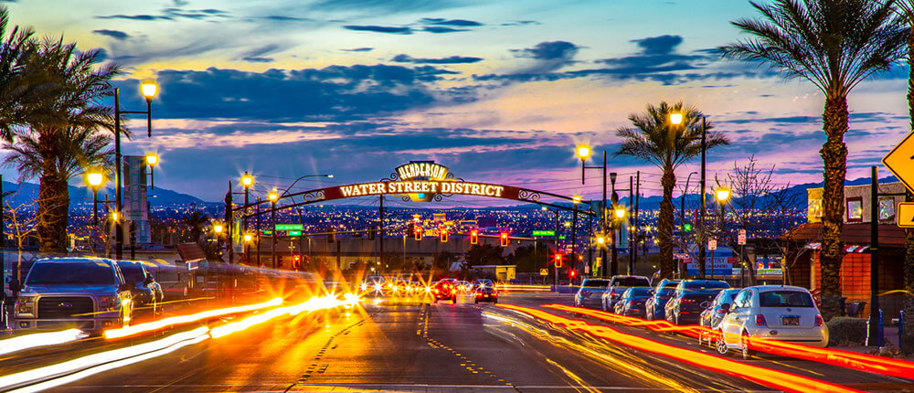 Street scene at dusk in Henderson's Water Street District. Pink clouds at sunset, and lit city in the background. Cars create light trails on the roads.
