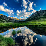 A lush green mountain scene with white puffy clouds in a dark blue sky, reflected in a foreground pond.