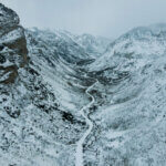 An aerial shot of a snowy mountain pass. A snow-covered road winds through the valley.