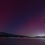 Subtle aurora borealis lights the starry sky with pinks and purples, reflected in a body of water with lit buildings and traffic surrounding.