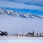 A ranch house with windmill and trees covered in hoarfrost, with low clouds and snowy mountains in the background.