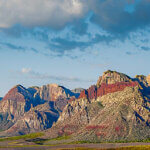 A mountain range with bands of red rock running through it. Green valley in the foreground ad puffy white clouds with blue skies above.