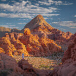 A person stands on the edge of a valley among tall red rocks. White clouds in a blue sky above.